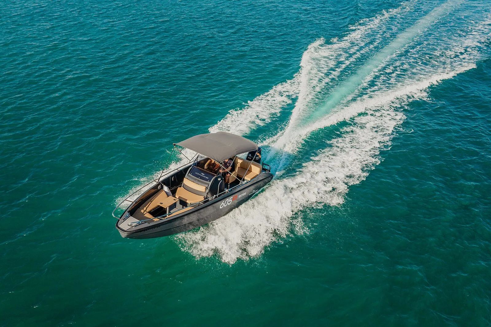 Aerial view of the OceanMaster 605, a black motorboat with a grey bimini top, cruising on turquoise water